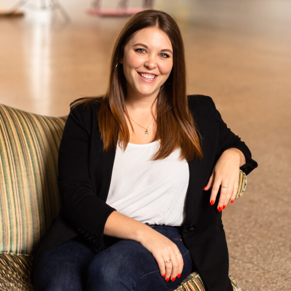 Women in a white blouse and black blazer sitting on a green and brown striped couch smiling at the camera