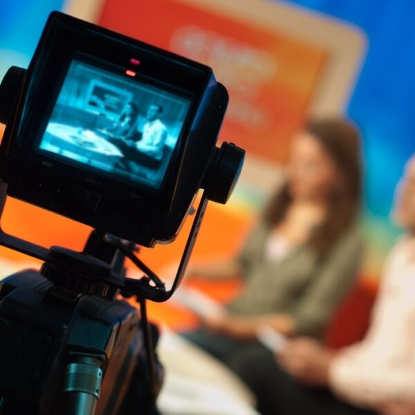 Two news anchors sitting on a couch with a camera in front of them