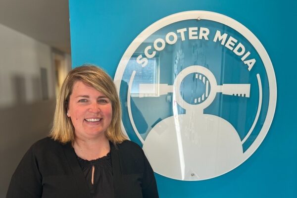 Danielle Reynolds, Public Relations Account Supervisor, standing in front of a blue wall with the Scooter Media logo.
