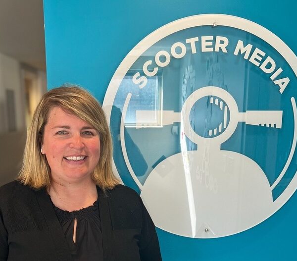 Danielle Reynolds, Public Relations Account Supervisor, standing in front of a blue wall with the Scooter Media logo.