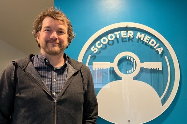 Pat LaFleur, Public Relations Manager, standing in front of a blue wall with the Scooter Media logo.