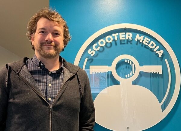 Pat LaFleur, Public Relations Manager, standing in front of a blue wall with the Scooter Media logo.